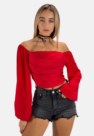 Top corto rojo sin mangas con detalles fruncidos y mangas largas acampanadas, combinado con pantalones cortos de mezclilla negra deslavada. Acento de gargantilla negra.
