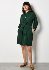 Grünes, knopfbares Kleid mit einem Bindeband vorne, langen Ärmeln, die an den Bündchen hochgerollt sind, und seitlichen Taschen. Glattes, leichtes Material.