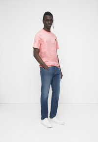 Κοντομάνικο ριγωτό μπλουζάκι σε ροζ και λευκό χρώμα με ένα μικρό γραφικό στο στήθος, συνδυασμένο με μπλε τζιν και λευκά αθλητικά παπούτσια.