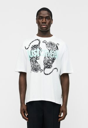 Čierny muž nosiaci bielu oversized tričko s dvoma čiernymi tigrami a svetlomodrým nápisom „Just Cavalli“, stojaci pred jednoduchým bielym pozadím.