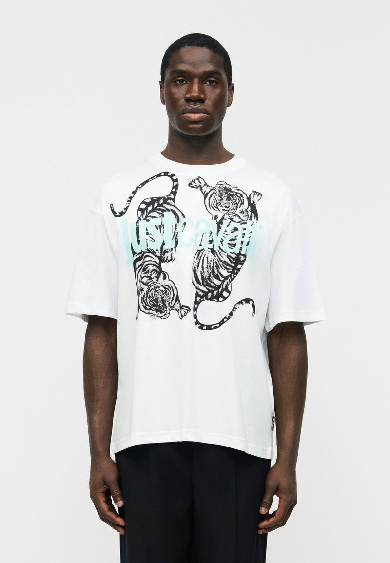 Μαύρος άνδρας που φοράει λευκό φαρδύ T-shirt με δύο μαύρες τίγρεις και ανοιχτό μπλε κείμενο "Just Cavalli", στέκεται μπροστά σε απλό λευκό φόντο.