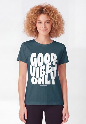 Junge Frau mit lockigem Haar, die ein dunkles T-Shirt mit großem weißen Text mit der Aufschrift „"GOOD VIBES ONLY"“ auf schlichtem Hintergrund trägt.