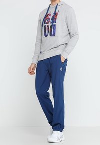 Grauer Hoodie mit buntem Grafikdruck und blauen Kordeln, kombiniert mit marineblauen Hosen und weißen Sneakers.