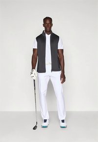 Σετ γκολφ με μαύρο αμάνικο γιλέκο, λευκό πουκάμισο με γιακά και λευκό παντελόνι. Λεπτομέρειες περιλαμβάνουν λευκή ζώνη και παπούτσια με μπλε λεπτομέρειες.