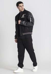 Čierna varsity bunda so šedými koženými rukávmi, s vyšitým písmenom "K", vyšívanými detailmi, prúžkovým lemom, skombinovaná s čiernymi nohavicami a bielymi teniskami.