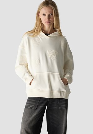 Blonde Frau trägt einen weißen Hoodie mit strukturierten Herzmustern und schwarze Jeans mit weitem Bein, Hände in den Taschen des Hoodies.