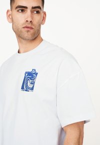 Camiseta blanca de algodón con un diseño gráfico azul en el pecho, con un corte holgado y mangas cortas.