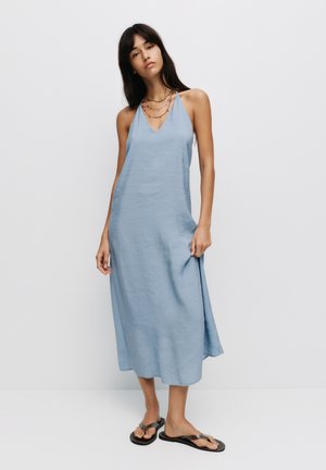 Robe midi sans manches, bleu clair, avec décolleté en V, texture douce, tissu fluide et fentes latérales. Portée avec des tongs noires.