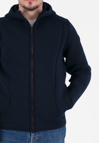 Felpa con zip di colore blu navy, dal design aderente, con una texture di tessuto liscia e chiusura frontale a zip. Presenta un cappuccio e tasche laterali.