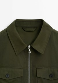 Veste vert olive avec col pointu, fermeture éclair argentée à l’avant et deux poches poitrine avec rabats boutonnés.