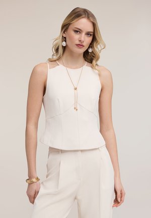 Blondynka w kremowym, bezrękawnikowym topie i dopasowanych spodniach, ozdobiona dużymi perłowymi kolczykami, złotymi bransoletkami oraz długim złotym naszyjnikiem.