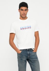 Lühikeste lokkis juustega mees, seljas valge 1985. aasta Tommy Jeans t-särk ja sinised teksad, käed taskutes, seismas valge tausta ees.