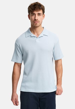 Mann mit kurzen lockigen Haaren, trägt ein hellblaues, kurzärmeliges Strick-Poloshirt und dunkle Hosen, steht vor einem einfachen Hintergrund.