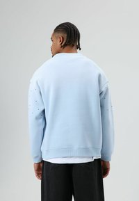 Sweat-shirt bleu clair à coupe décontractée ; présente des clous décoratifs sur les manches et des poignets côtelés. Associé à un pantalon foncé.