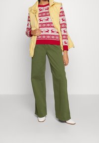 Persona che indossa un maglione con renna rosso e bianco, un gilet imbottito giallo senza maniche, pantaloni larghi verde oliva e scarpe bianche a punta.