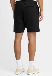 Pantaloni corti neri in cotone, con un elastico in vita, tasca posteriore e cuciture laterali. Abbinati a sneakers beige e calzini bianchi.