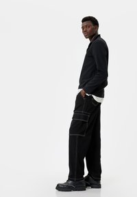 Zwarte langarmshirt met ribbeltextuur, gecombineerd met zwarte cargo-broeken met witte stiksels en zijzakken, en zwarte schoenen.
