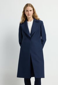Cappotto lungo blu navy con motivo a griglia testurizzato, ampi rever, chiusura a un bottone e tasche laterali. Indossato sopra una camicia bianca.
