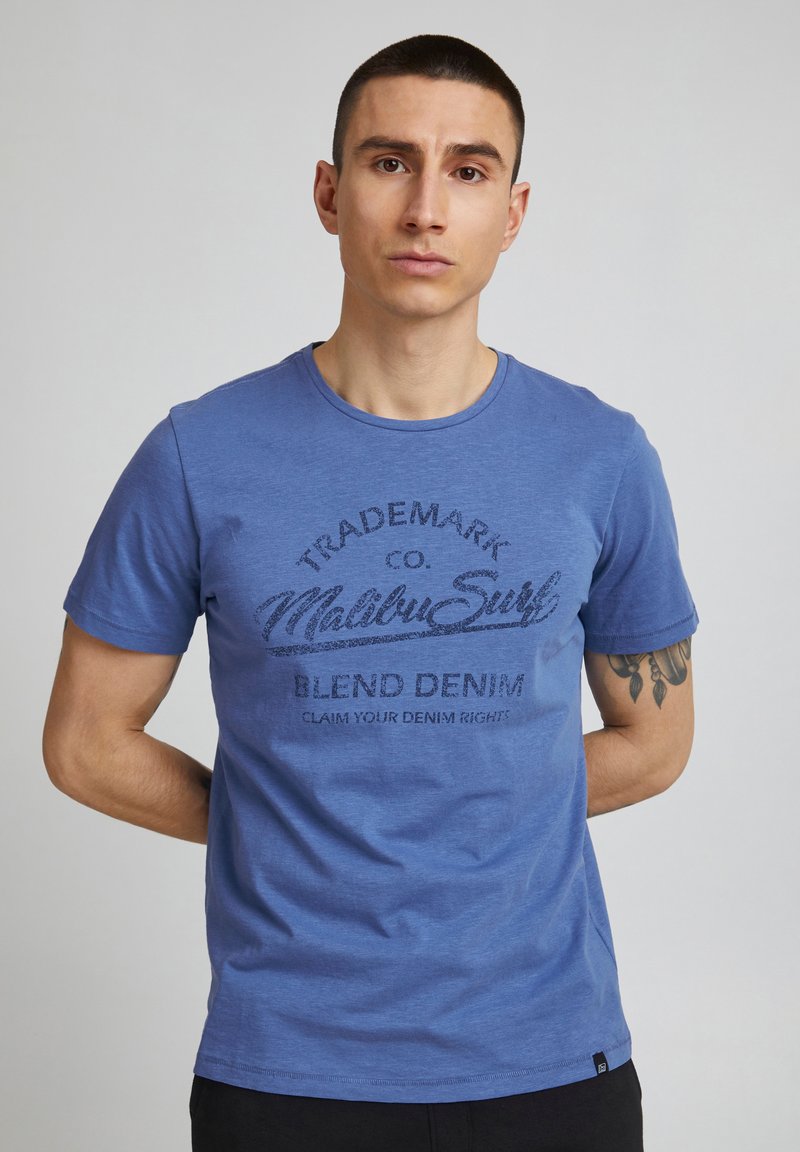 Muž s krátkymi vlasmi v modrom tričku "Malibu Surf Blend Denim" stojí s rukami za chrbtom na jednoliatom pozadí.
