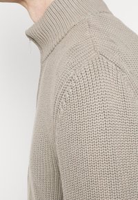 Ljusbeige stickad tröja med hög ribbad krage och halvv zipper-design; har texturerad sömnad och en åtsittande silhuett.