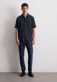 Korte mouwen, donkerblauwe button-up shirt van een gestructureerde stof, gecombineerd met strakke donkere jeans en zwarte veterschoenen.