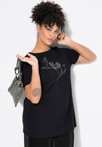 Camiseta negra con un diseño floral hecho de tachuelas plateadas. Modelo sosteniendo un bolso negro y plateado mientras muestra un tatuaje en su brazo.