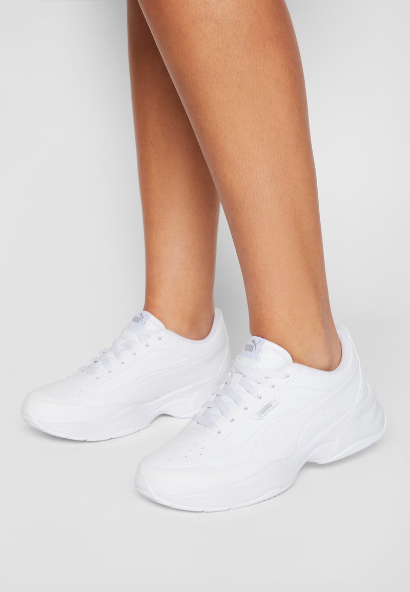 Baskets basses blanches avec semelles épaisses portées pieds nus sur un fond clair uni.