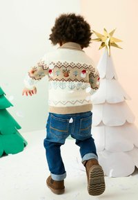 Mėlynos džinsinės kelnės ir rudų aulinukų derinys su mezginiu kreminės spalvos megztiniu, puoštu šventiniais motyvais, įskaitant elnius ir sniego senelius.