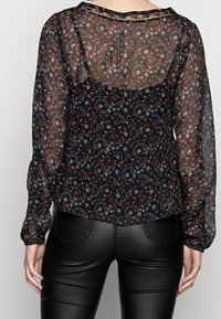 Svart blus i genomskinligt tyg med paisley- och blommönster i röd, blå och vit. Har långa ärmar och en avslappnad passform.