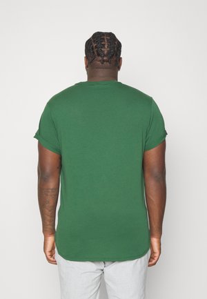 Homme aux cheveux tressés portant une chemise verte à manches courtes et un pantalon gris clair, debout de dos contre un fond uni.