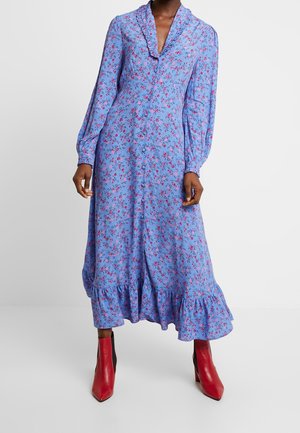 Robe à manches longues, bleue à motifs floraux avec un ourlet volanté, ornée de fleurs rouges et roses. Stylisée avec des bottines à talons rouges et noires.