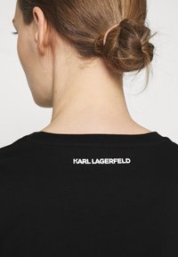 Černé tričko s kulatým výstřihem, vyrobené z hladkého materiálu, s bílým logem "KARL LAGERFELD" vytištěným na horní části zad.