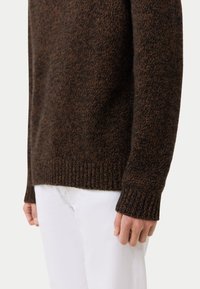 Brauner Strickpullover mit strukturiertem Muster, gerippten Bündchen und Saum. Kombiniert mit weißen Hosen, die einen figurschmeichelnden Schnitt zeigen.