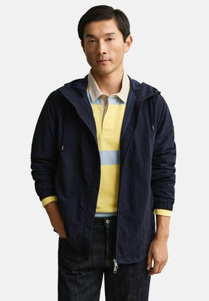 Homme portant une veste à capuche bleu marine par-dessus un polo à rayures jaunes et bleu clair, et un jean foncé, debout avec une main dans la poche.