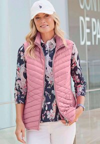 Helles rosa, gestepptes Weste mit Reißverschluss über einem marineblauen Blumenshirt. Das Model trägt eine weiße Kappe, die einfache Designs und Texturen zur Schau stellt.