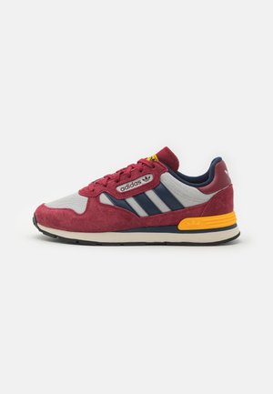 Baskets Adidas rouges et grises avec des rayures bleu marine, détail jaune au talon et semelle noire sur fond blanc, vue de côté.