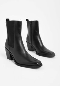 Stivali alla caviglia in pelle nera con punta quadrata, pannelli laterali elasticizzati, chiusura con zip e un tacco block moderato. Texture liscia con un design minimale.