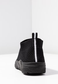 Chaussures noires tricotées à enfiler, présentant une texture en maille, une bande blanche à l'arrière, et une semelle en caoutchouc avec une semelle à motif.