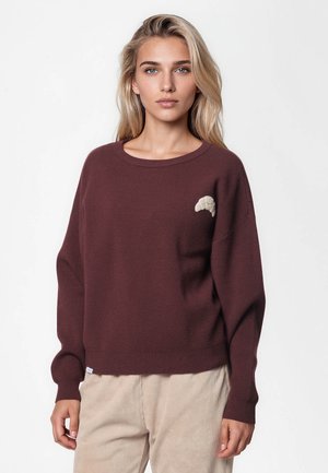 Jeune femme aux longs cheveux blonds porte un pull marron foncé avec un petit motif croissant beige près de l'épaule gauche et un pantalon beige.