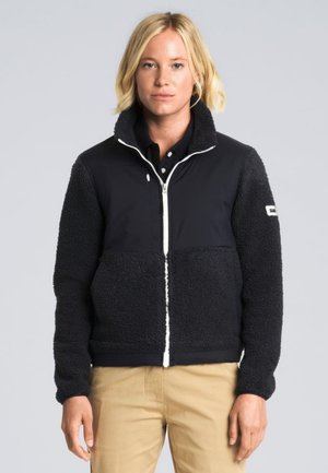 Modelo rubio con una chaqueta de forro polar negra con cremallera frontal blanca y pantalones beige, de pie sobre un fondo gris claro.