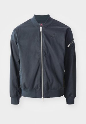 Giubbotto bomber blu scuro con zip frontale, polsini e collo a costine. Presenta un logo ricamato sul petto e una tasca con zip sulla manica.