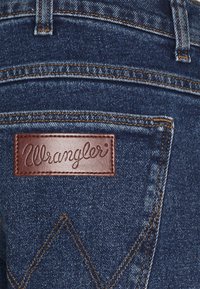 Jeans in denim blu scuro con una toppa in pelle marrone con il logo "Wrangler" impresso, cuciture a contrasto e design classico delle tasche.
