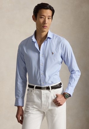 Férfi világoskék csíkos, gombos ingben, fehér nadrágban, fekete övvel és zöld karórával, semleges háttér előtt áll.