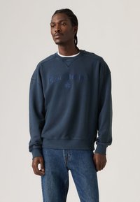 Μπλε σκούρο φούτερ με στρογγυλή λαιμόκοψη, κεντημένο λογότυπο «Levi Strauss» και ριμπ μάτια στα μανίκια, σε συνδυασμό με μπλε τζιν παντελόνι.