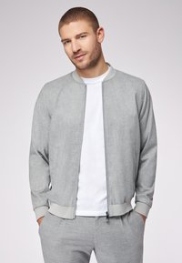 Giacca bomber grigia con finitura testurizzata, zip frontale, colletto e polsini a costina, abbinata a una T-shirt bianca e pantaloni a quadri grigi.