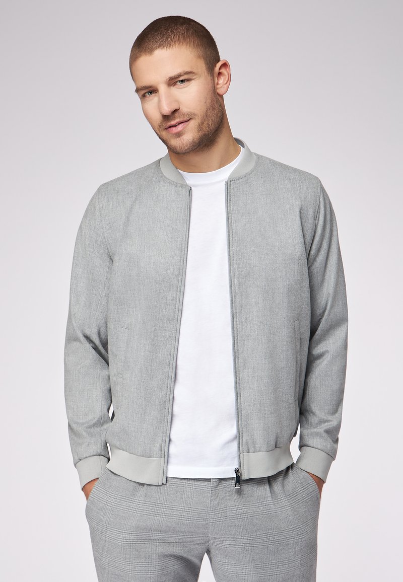 Giacca bomber grigia con finitura testurizzata, zip frontale, colletto e polsini a costina, abbinata a una T-shirt bianca e pantaloni a quadri grigi.