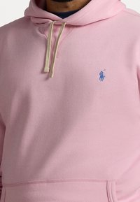 Rosa hoodie i mjukt tyg, med en framficka, beige dragskor och en ljusblå logotyp på vänster bröst.
