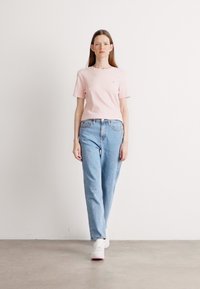 Chemise rose clair à manches courtes associée à un jean droit bleu clair. Le modèle porte des baskets blanches. Fond minimaliste.