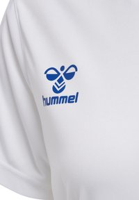 Camiseta atlética blanca con el logo "hummel" en azul y un diseño gráfico en la parte superior izquierda. Tejido suave y liso con un ligero brillo.