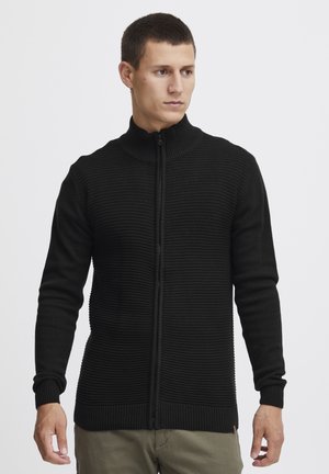 Svart stickad cardigan med hög krage och full dragkedja. Har texturerad ribbstickning och åtsittande muddar, tillverkad av ett mjukt material.